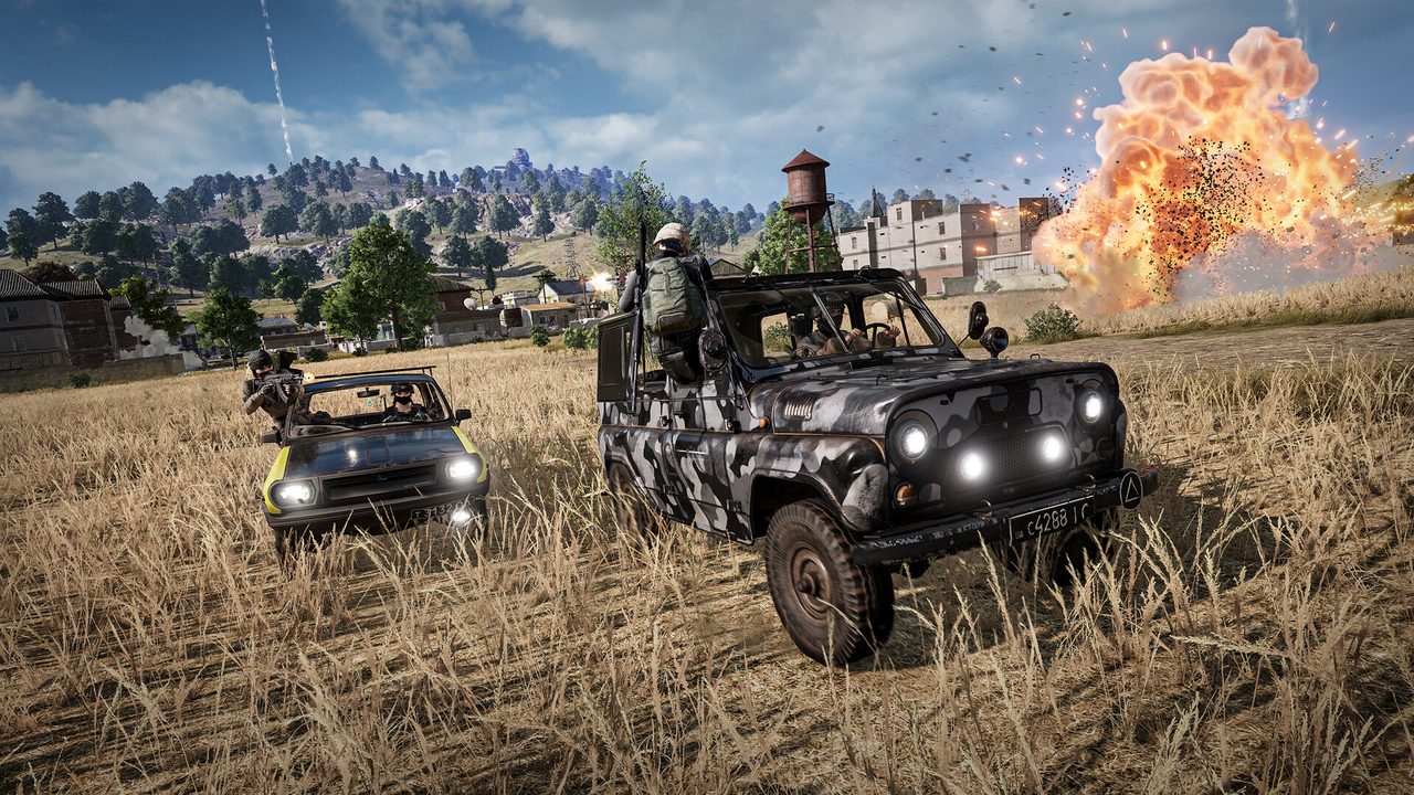 Two open-top jeeps carrying armed players speeding across a golden grass field as a large explosion erupts near a water tower in the background.